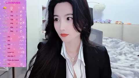 Media: Video of a slender East Asian woman with long black hair, fair skin, and red lipstick, wearing a black blazer over a white shirt. Background includes a bed with gray bedding, a pink plant, and a digital clock.