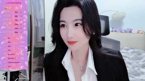 Media: A video of an East Asian woman with fair skin, long black hair, and red lipstick, wearing a black blazer over a white shirt, in a modern bedroom with a bed and wall-mounted TV.