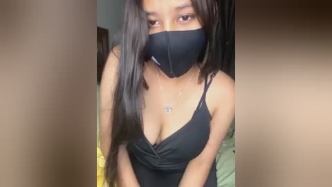 Media: Video of a young woman with medium skin tone, long dark hair, wearing a black face mask, black spaghetti strap dress, and a butterfly necklace, indoors.