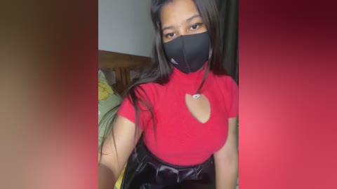 Media: Video of a young Asian woman with medium skin tone, wearing a red turtleneck, black mask, and black skirt, seated indoors with a wooden headboard and yellow-green blanket in the background.