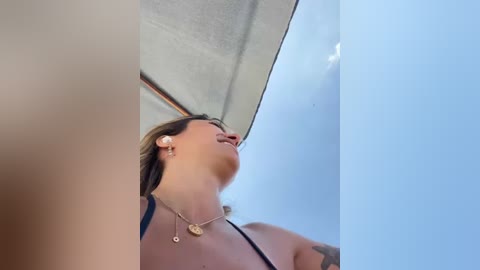 Media: Video of a woman with light skin, brown hair, and a tattoo on her arm, wearing a black bikini top, smiling up towards a clear blue sky, with a sunlit, reflective surface behind her.