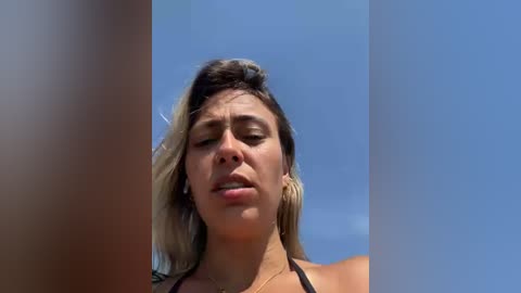 Media: Video of a woman with light skin and shoulder-length, blonde hair, wearing a black bikini top, squinting against the bright blue sky.