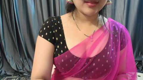 Media: Video of a smiling woman with fair skin, wearing a black blouse with white polka dots and a semi-transparent pink sari with small white dots. She has black hair tied back, and the background features dark, flowing fabric.