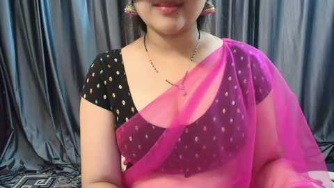 Media: Video of a smiling woman with light skin, wearing a black blouse with white polka dots, draped in a vibrant pink saree with a sheer drape, set against a grey, patterned backdrop.