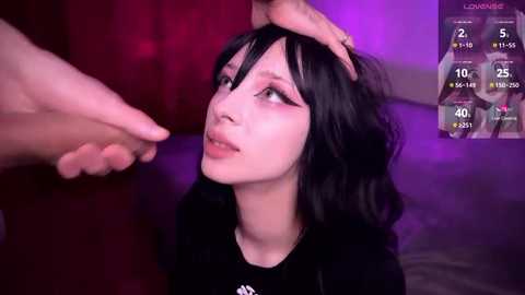 Video of a pale-skinned woman with dark hair and eyeliner, sitting with her head in another person's hand. Background has purple and red lighting.