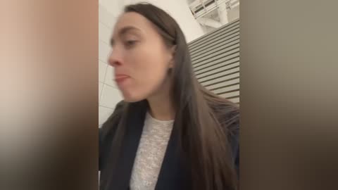 Media: A blurry video of a young woman with long brown hair, wearing a dark jacket over a light grey top, in a bathroom with white tiles and a striped wall.