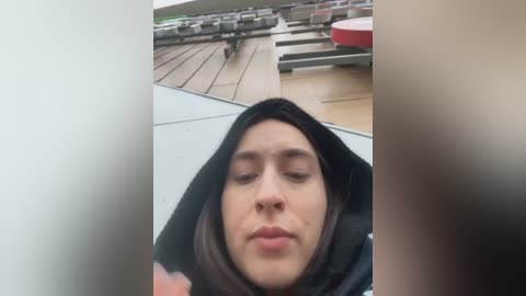 Media: Video of a woman in a black hijab, eyes closed, seated outdoors on a wooden deck with bleachers in the background.