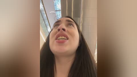 Media: Video of a smiling young woman with long dark hair, fair skin, and a pleasant expression, taken from a low angle, showing her head and upper torso. Background features modern, minimalistic interior design with glass partitions and light fixtures.