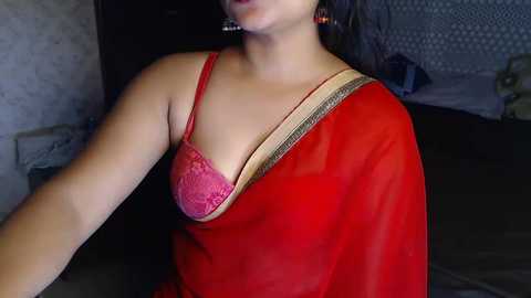 Media: Video of a South Asian woman with medium skin tone, wearing a red sari and a pink lace bra, partially visible, in a dimly lit room.