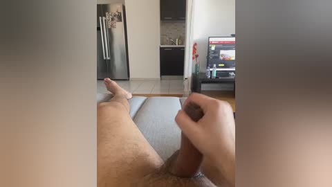 Media: Video of a man lying on a bed with his legs spread, his hand holding his erect penis. Background shows a modern kitchen with a refrigerator, cabinets, and a TV.