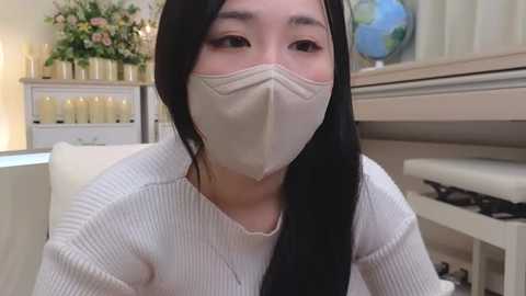 Media: Video of an Asian woman with long black hair, wearing a white ribbed sweater and a beige mask, seated indoors with a floral arrangement and a globe in the background.