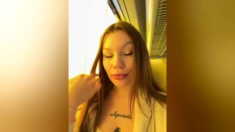 Media: Video of a young woman with long brown hair, eyes closed, and a tattoo on her chest, sitting in a car with golden-yellow lighting.