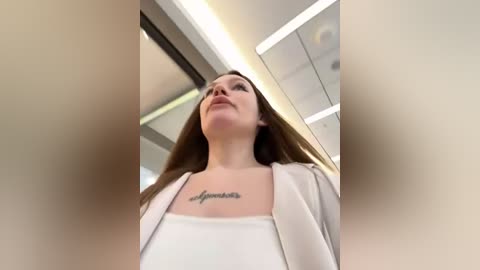 Media: Video of a young woman with long brown hair, wearing a white blouse, standing inside a modern, brightly lit room with large windows and reflective surfaces.