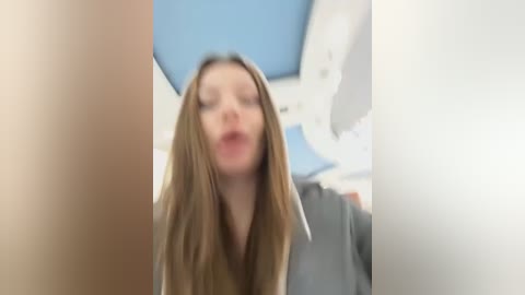 Media: A blurry, out-of-focus video of a young woman with long, straight, light brown hair, wearing a gray jacket, standing in a brightly lit room with blue and white ceiling tiles.