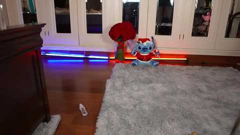 Media: Video of a cozy living room with plush toys, including a plush Lilo and Stitch and a plush red rose, on a light gray shaggy rug. The background features white cabinets with glass doors reflecting the colorful LED lights underneath.