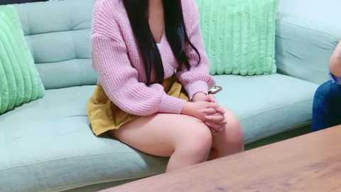 Media: Video of a young woman with long black hair, wearing a pink knitted cardigan, yellow skirt, and white shirt, sitting on a light grey couch with green pillows.