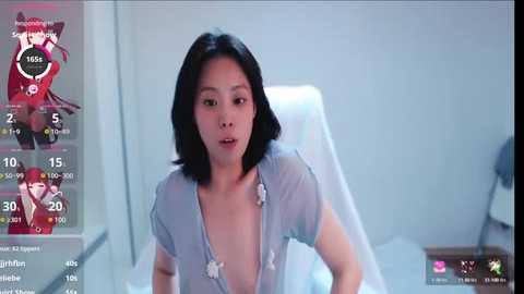 Media: A video of an East Asian woman with black hair, wearing a light blue shirt, in a sterile room, with a game UI overlay.