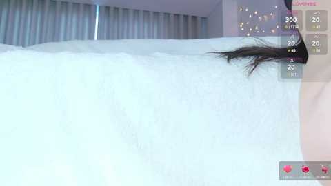 Media: Video of a person with long black hair lying on a white bed with a soft, fluffy texture. The background shows a modern bedroom with light-colored curtains and a digital interface displaying the time, 3000, and \"Xtra.\