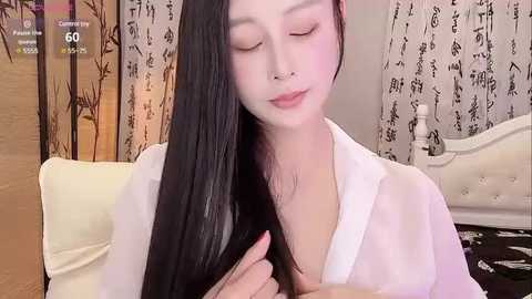 Media: Video of a young East Asian woman with long black hair, wearing a sheer white blouse, sitting in a room with patterned curtains and a white bed.