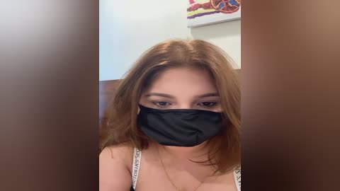 Media: A video of a young woman with fair skin and shoulder-length auburn hair, wearing a black mask, white top, and a necklace, standing in a bathroom with brown walls and colorful artwork.