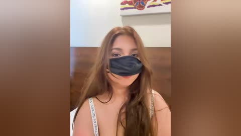 A video of a young woman with long, straight brown hair, wearing a black face mask, white top, and a white purse. The background shows a wooden wall with abstract art.