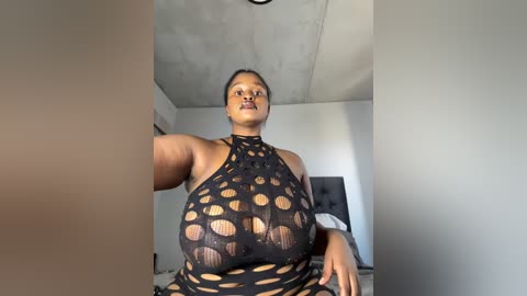 Media: A video of a curvy Black woman with dark skin and braided hair, wearing a revealing black halter top with large cut-outs, seated in a modern, minimalist room with grey walls and a ceiling fan.