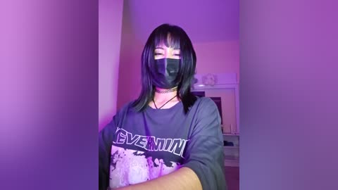 Video of a young Asian woman with long black hair, wearing a black face mask and a dark, patterned \"SVEENI\" t-shirt, standing indoors with purple lighting.