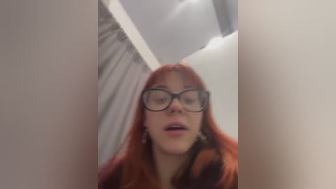 Media: Video of a young woman with long, fiery red hair, wearing black-framed glasses, standing in a narrow, dimly lit hallway with white walls and a ceiling light.