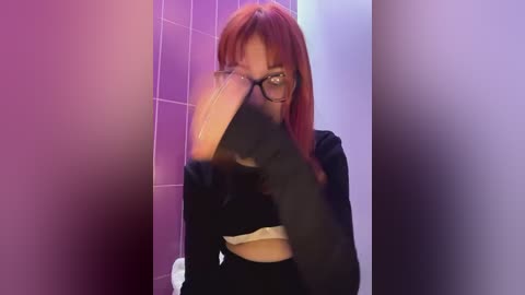 Media: Video of a woman with long red hair, wearing black glasses and a black top, hiding her face with her left hand, standing in a dimly lit restroom with purple-tiled walls.