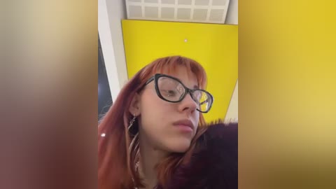 Media: Video of a woman with long red hair and glasses, wearing a dark outfit, in a yellow and white room with a tiled ceiling.