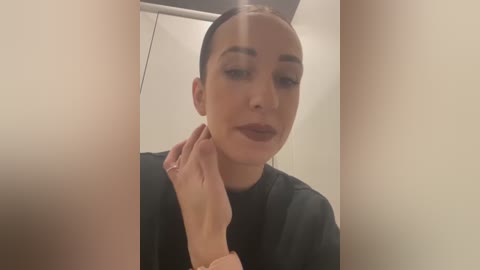 Media: A video of a woman with fair skin, dark hair, and light makeup, wearing a black top, touching her neck, in a dimly lit room with beige walls.