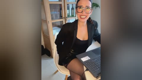 Media: Video of a smiling woman with glasses, wearing a black blazer and black skirt, sitting at a white desk with a laptop, in a modern office with wooden cabinets and a plant.