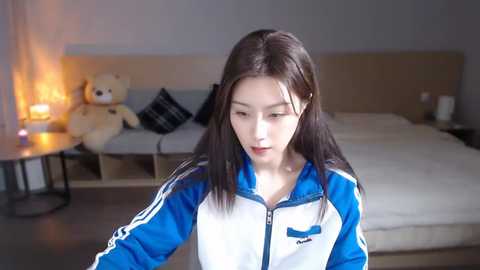 Media: Video of an East Asian woman with long black hair, wearing a blue and white athletic jacket, standing in a modern bedroom with a bed, teddy bears, and a lit lamp.