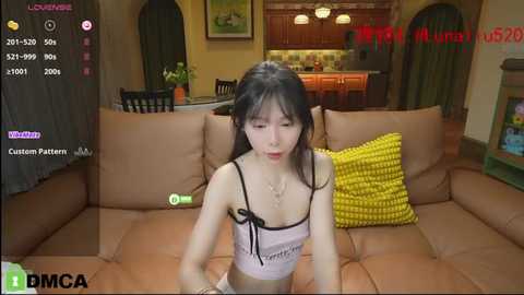 Media: Video of an Asian woman with fair skin, wearing a white lingerie top, sitting on a brown couch in a modern kitchen.