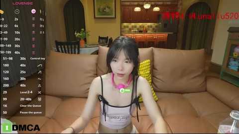 Media: Video of an Asian woman with long black hair, wearing a pink necklace, seated on a brown couch in a cozy living room with wooden furniture.