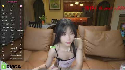 Media: Video of an East Asian woman with black hair, wearing a pink lace top, sitting on a brown couch in a modern living room.