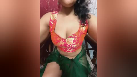 Media: Video of a South Asian woman with medium skin tone, wearing a red and gold floral bra and green sheer sarong, seated indoors against a floral patterned wall.