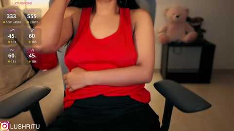 Media: Video of a woman in a red tank top, sitting in a chair, with a teddy bear and a TV in the background.