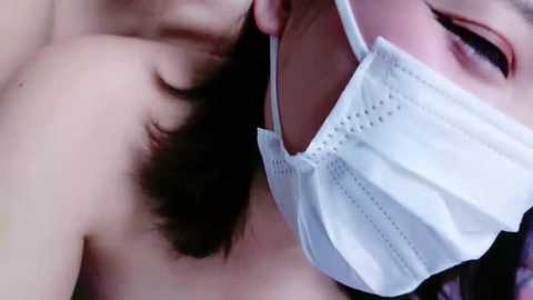 Media: Video of a close-up of a person with a white surgical mask partially covering their face, revealing dark hair and a hint of a bare shoulder, suggesting intimacy.