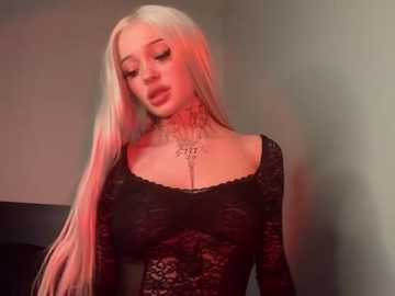 Media: Video of a young, light-skinned woman with long platinum blonde hair, wearing a black lace bodysuit. She has tattoos on her neck and collarbone. The background is dimly lit with a red glow, creating a moody atmosphere.