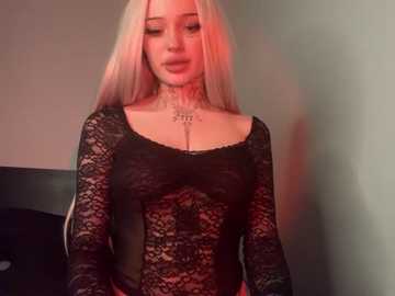 Media: Video of a young woman with long platinum blonde hair, fair skin, and a slender physique, wearing a black lace bodysuit. She stands against a plain grey wall, bathed in warm, red-toned lighting.