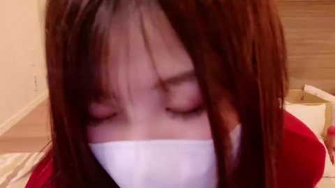 Media: A close-up video of a woman with long brown hair wearing a white mask and a red jacket, seated on a wooden floor. The background shows a beige wall and a cardboard box.