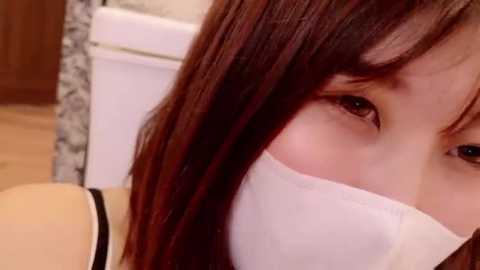 Media: Video of a young Asian woman with brown hair, wearing a white surgical mask, looking to the side, taken indoors in a bathroom with a white toilet and tiled walls.