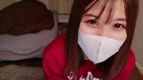 Media: Video of a young Asian woman with long brown hair, wearing a red hoodie and white mask, indoors on a wooden floor, next to a grey couch and a brown pillow.