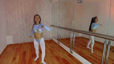 Media: A video of a joyful Asian woman with long black hair in a light blue crop top and leggings, dancing on a wooden floor in a mirrored ballet studio.