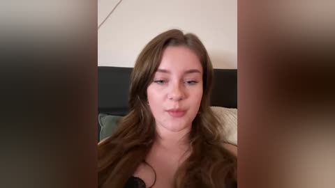 Media: A video of a young woman with long brown hair, wearing a black bra, sitting on a bed with beige and green pillows in the background.