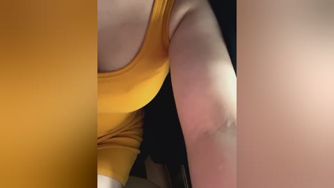 Media: Video of a close-up, side view of a person's arm and torso in a mustard yellow tank top. The skin is fair and smooth, with a small bruise on the upper arm. The background is dark.