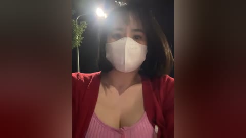 Media: Video of a woman with medium-length brown hair, wearing a red cardigan over a pink tank top, a white face mask, and a street lamp in the background.