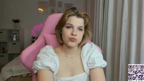 Media: Video of a young Caucasian woman with wavy blonde hair, fair skin, and blue eyes, wearing a white, short-sleeved top, sitting in a pink gaming chair, indoors with light walls, white curtains, and a white shelving unit.