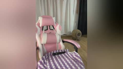 Media: Video of a pink and white gaming chair with adjustable armrests and backrest in a cozy room with a furry cat toy and white curtains.